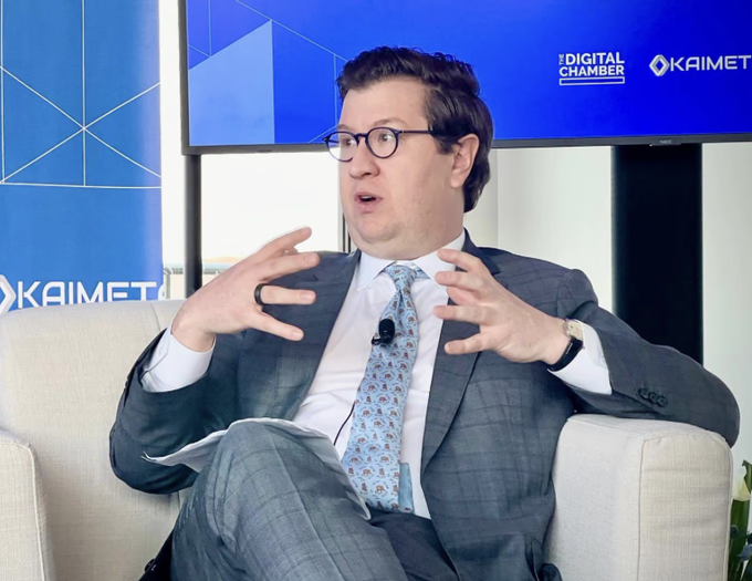 A man in a gray suit and glasses gestures animatedly while seated in a chair at what appears to be an industry event, with "KAIMET" and "THE DIGITAL CHAMBER" logos visible on the blue backdrop behind him. The image relates to the post as it may depict Tyler Williams, the U.S. Treasury digital asset policy chief mentioned in the headline, in a typical speaking engagement setting related to crypto policy.