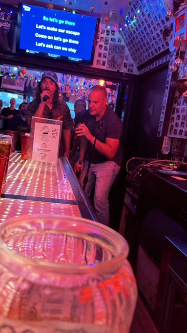 Two people sing karaoke at a bar with colorful lights, while lyrics including "So let's go there" and "Let's make our escape" appear on a screen above them.