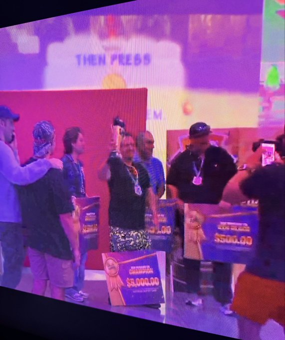 A person holds up a trophy on a brightly lit stage, surrounded by others, with visible prize money signs for '$8,000.00' and '$3,500.00' beneath a screen reading 'THEN PRESS'.