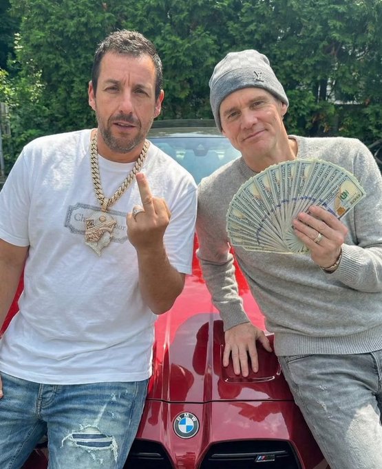 Adam Sandler (right) and co-star Mark Strong pose in front of a red BMW, with Sandler flashing a fan of $100 bills in a style reminiscent of his crypto-promoting meme, tying into the "$Lloyd" Solana token reference.