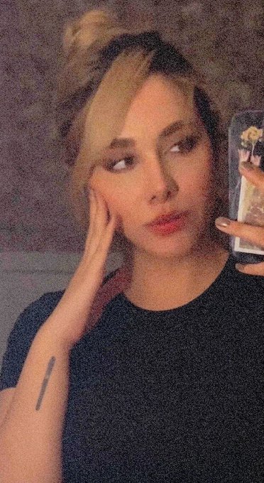 A grainy selfie of a young woman with blonde hair styled in two buns, wearing a black t-shirt and posing thoughtfully while holding a phone with a yellow floral case.
