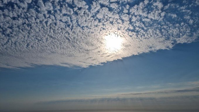 A scenic view of the sun shining brightly through a layer of patterned clouds against a deep blue sky, with a distant horizon visible at the bottom.