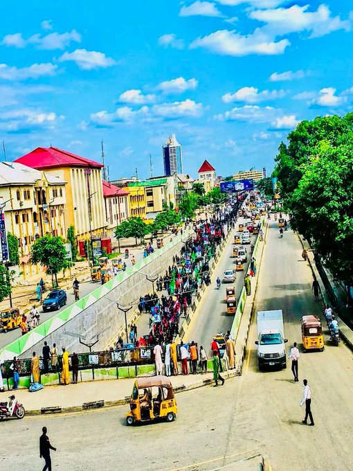 An aerial view of a bustling city street with pedestrians, yellow auto-rickshaws (kekes), cars, and colorful buildings under a bright blue sky with scattered clouds.