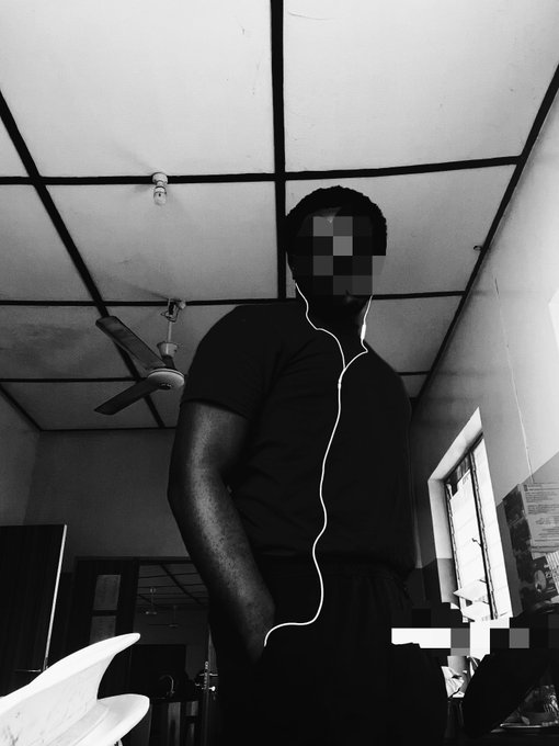 A black-and-white photo of a man in a dark t-shirt standing in a room with a ceiling fan, wearing wired earphones, with his face blurred out for privacy.