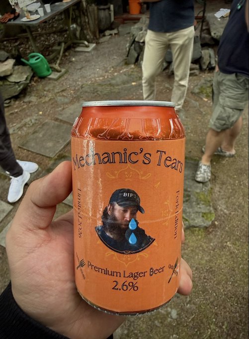 A hand holds up an orange can of 'Mechanic's Tears Premium Lager Beer' at 2.6% ABV, featuring a logo of a bearded man with a blue teardrop, set against a background of a stone patio with people.