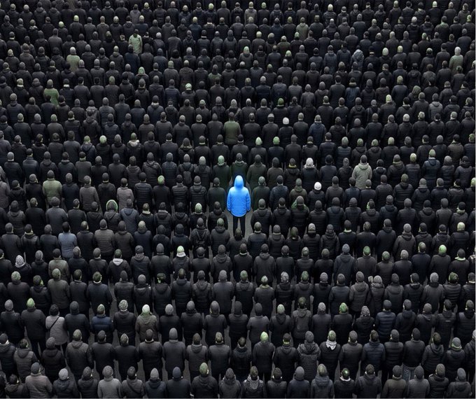 An overhead view of a dense crowd of identical gray figures with one bright blue figure standing out in the center, symbolizing individuality or standing out from the masses.