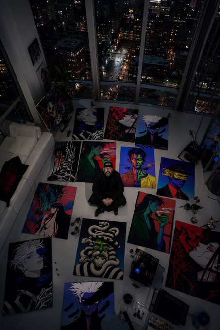 A man sits cross-legged on the floor of a high-rise apartment surrounded by his colorful, pop-art style paintings, with a nighttime cityscape visible through the large windows behind him.