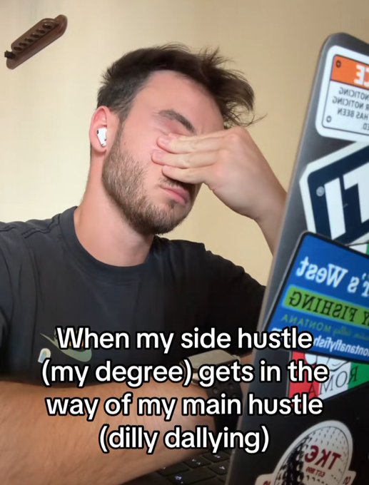 A man covering his face with his hand in frustration while looking at a laptop covered in stickers, with overlaid text reading 'When my side hustle (my degree) gets in the way of my main hustle (dilly dallying)'.
