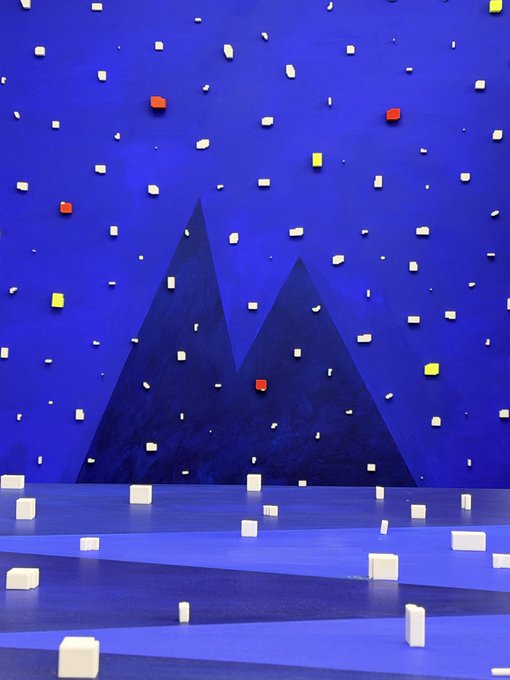An immersive art installation with deep blue walls and floor featuring dark triangular mountain shapes, scattered with small white, red, and yellow rectangular blocks mounted on the surfaces and resting on the floor.