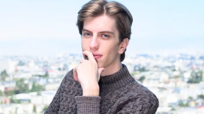 A young man with brown hair wearing a dark gray knit sweater poses thoughtfully with his hand near his chin against a blurred cityscape background.