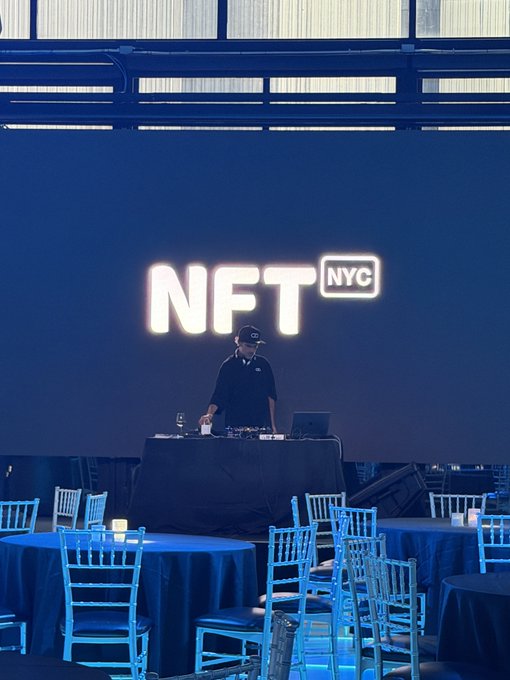 A DJ wearing a cap performs behind a table at an event, with a large illuminated 'NFT NYC' sign displayed on the blue wall behind him.