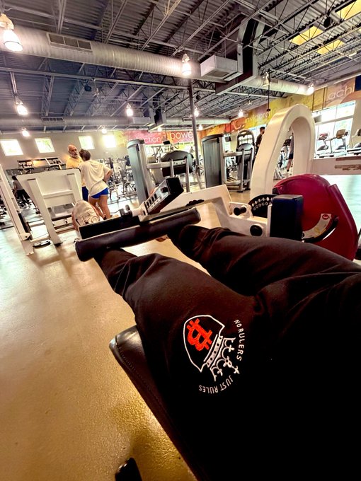 A first-person POV shot at a gym shows someone seated at a leg machine gripping the handles, wearing black sweatpants featuring a "Rutgers HODLERS" logo with a Bitcoin-style symbol. The image humorously ties the "Leg days are better when you Hodl Bitcoin" caption to the visible crypto-themed apparel, blending fitness with a playful nod to crypto culture.