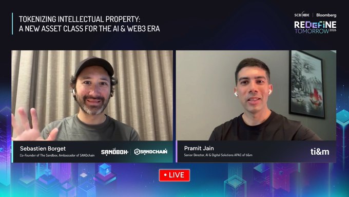 The image shows a screen with two men talking about tokenizing intellectual property, specifically focusing on the AI & Web3 era. The first man is wearing a gray shirt while the second man has dark hair and is wearing a black shirt. They are both smiling as they engage in conversation. In the background of the image, there is a picture hanging on the wall that adds to the overall atmosphere of the scene.