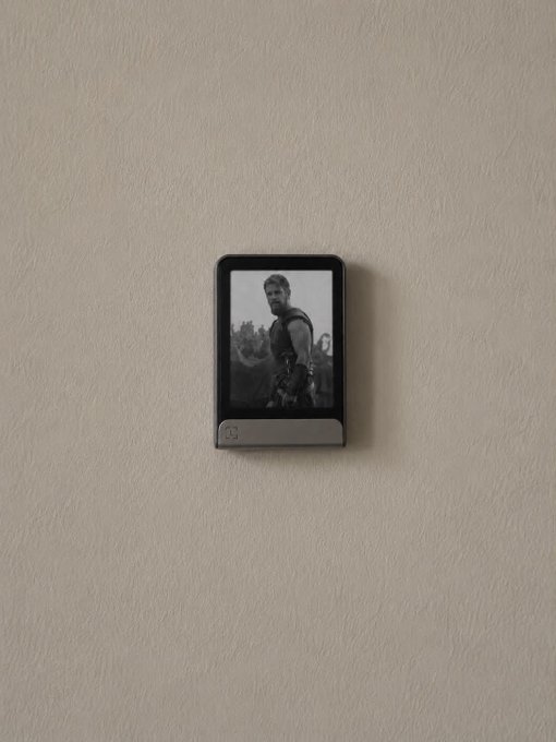 A small electronic photo frame mounted on a beige wall displaying a black-and-white image of a bearded warrior character, likely Thor.