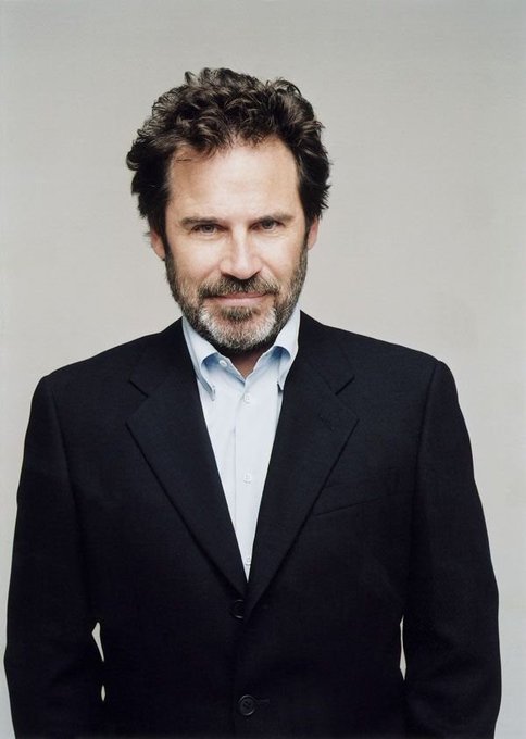 Professional portrait of a middle-aged man with curly brown hair and a beard, wearing a dark suit jacket over a light blue shirt, photographed against a gray background.