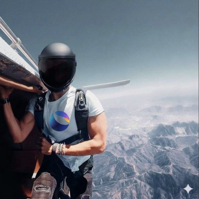 A person wearing a helmet and a harness with a colorful logo stands at the open door of a small aircraft high above a mountainous landscape.