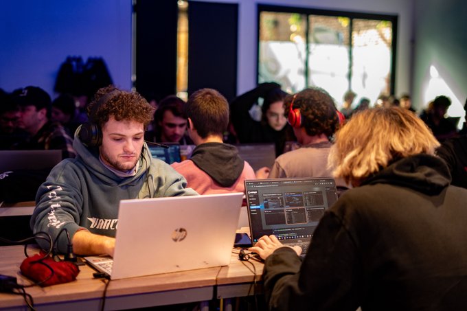 A group of focused developers wearing headphones work on laptops and code in a dimly lit room, suggesting a collaborative coding or hackathon environment.