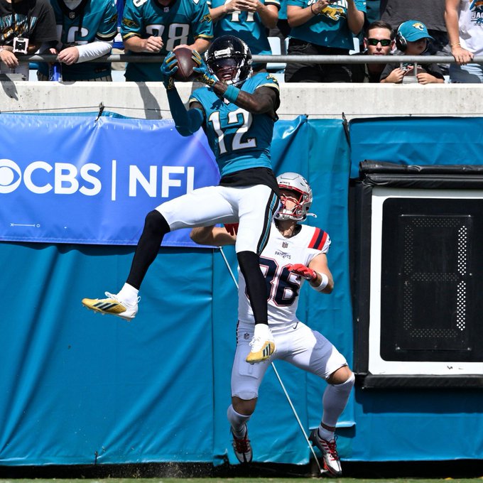A Jacksonville Jaguars wide receiver wearing jersey #12 leaps high to make a catch over a New England Patriots defender (#26) near the sideline during a game broadcast on CBS. The caption "CB1 !!! Finally man" celebrates the cornerback's interception, contrasting with the spectacular leaping catch shown in the image.