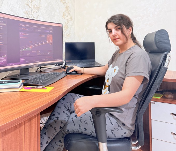 A young woman with dark hair sits in a black office chair at a wooden desk, using a mouse to look at a stock trading chart on her large monitor, with a laptop and notebooks nearby.