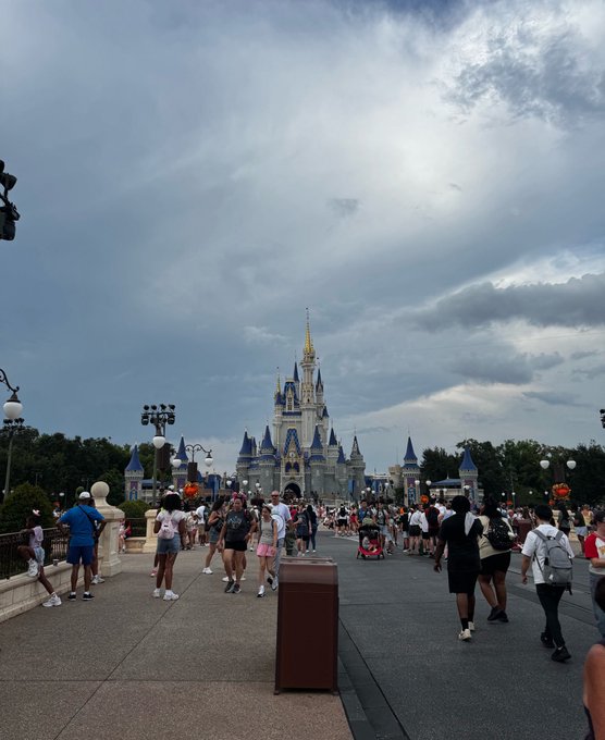Photograph of Cinderella Castle at Disney World viewed from Main Street, with crowds of tourists walking along the pathway under a cloudy sky.