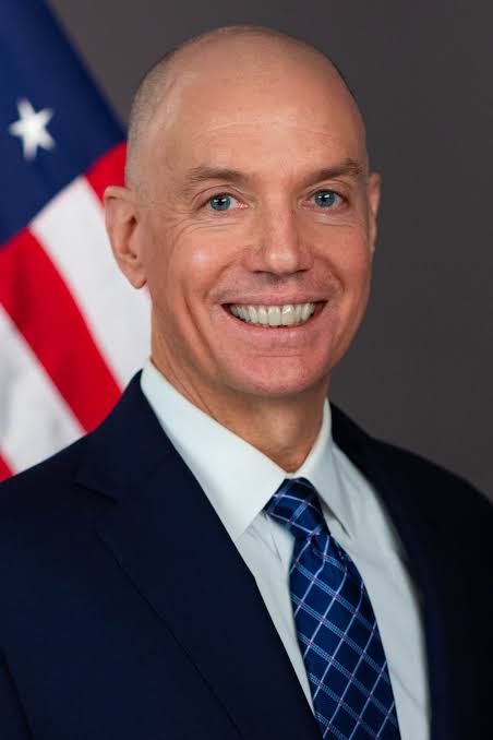 A professional portrait shows a smiling, bald man in a dark navy suit with a blue plaid tie, photographed against a gray background with an American flag visible on the left side. The image is associated with the announcement that SEC Principal Deputy Director of Enforcement Sam Waldon will depart the agency on July 31 after 14+ years of service.
