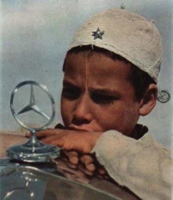 A young boy wearing a white cap with a star gazes longingly at a Mercedes-Benz hood ornament, his tender hands resting on the car, embodying the universal "Dream" of one day owning such a symbol of luxury and success.