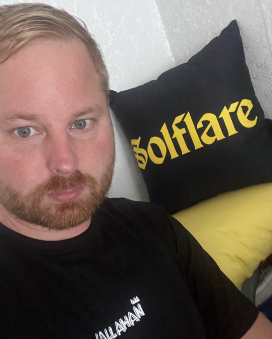 A close-up selfie of a blonde, bearded man wearing a black t-shirt with partially visible white text reading 'ALLAHAN', posing next to a black pillow with the yellow 'Solflare' logo.