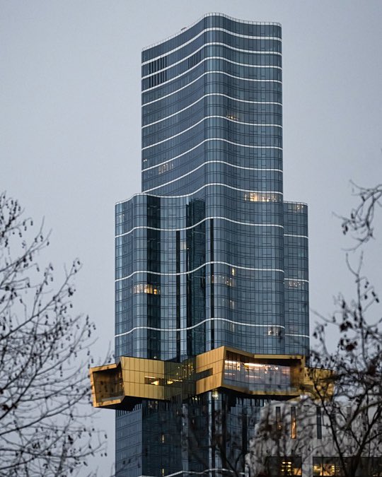 A tall, modern glass skyscraper with a distinctive wavy, tiered design and a golden protruding section near the bottom is photographed against a gray sky, framed by bare tree branches.