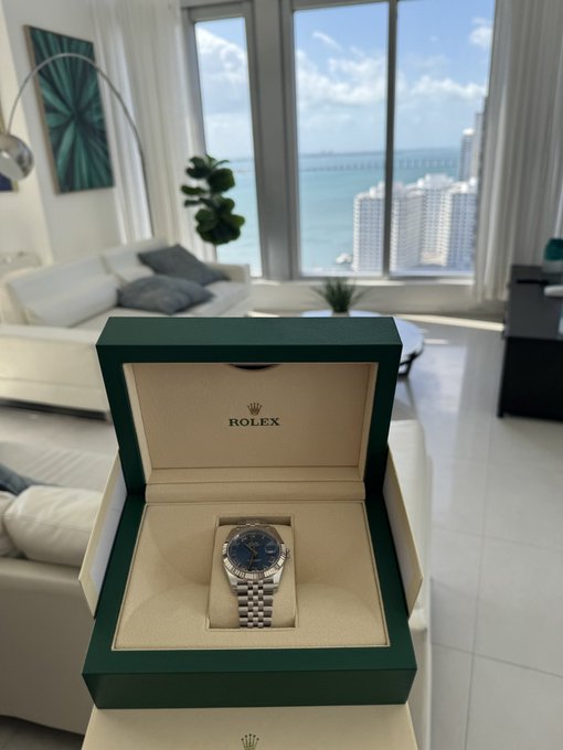 A Rolex watch with a blue dial and silver jubilee bracelet displayed in its green presentation box, set against a backdrop of a luxury apartment with an ocean view.