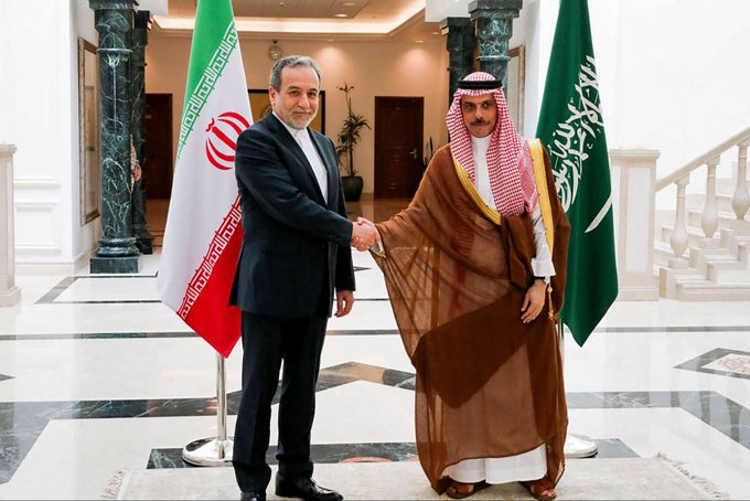 Two men shake hands in a formal setting between the Iranian and Saudi Arabian flags, with one wearing traditional Saudi robes and the other in a dark suit.