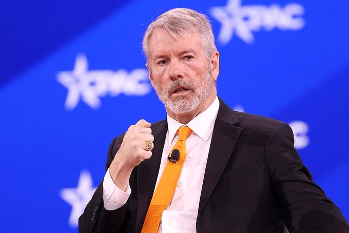 A gray-haired man with a beard, wearing a black suit and orange tie, gestures with a clenched fist while speaking at what appears to be a CPAC event (visible by the star logo backdrop).

The image relates to the post as it likely depicts Michael Saylor, the Strategy (formerly MicroStrategy) executive chairman, who the article questions about his decision to pause Bitcoin acquisitions and build cash reserves instead.