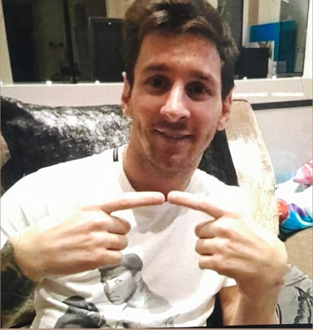 A selfie of soccer player Lionel Messi pointing at his chin and smiling, wearing a white t-shirt with a graphic print.