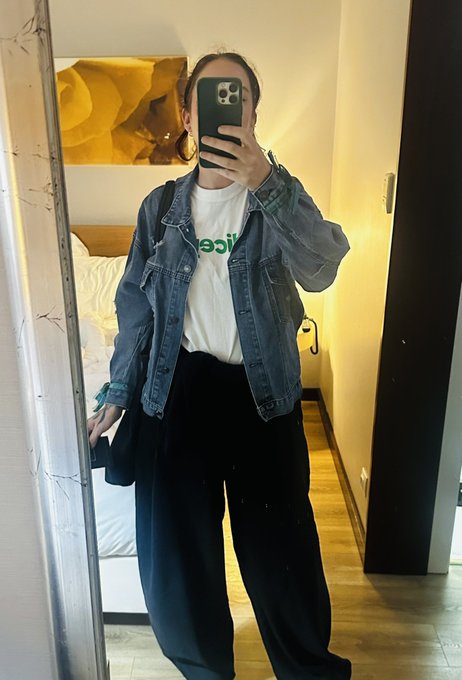 A mirror selfie of a person wearing an oversized denim jacket over a white t-shirt with green text and black pants, holding a dark smartphone.
