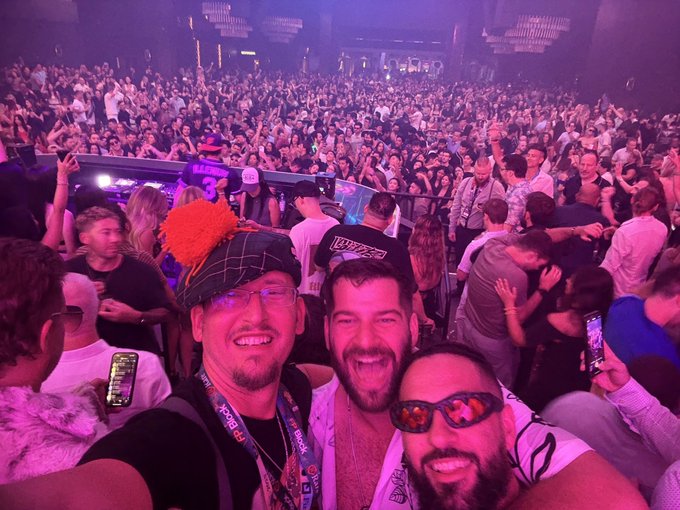 A large, energetic crowd fills a nightclub or concert venue under purple lighting, with three men in the foreground taking a selfie, one wearing a fuzzy orange and black hat.