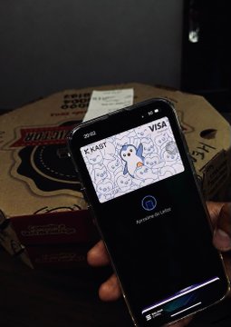 A hand holds an iPhone displaying a digital KTAP Visa card with a blue bird mascot illustration, positioned in front of a Pizza Hut box.