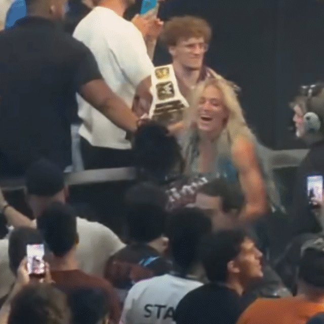 The image shows a group of people gathered around, with one person holding up an award. A woman wearing a white shirt is laughing while another person holds her trophy. The crowd appears to be enjoying the moment as they watch the winner celebrate her victory.