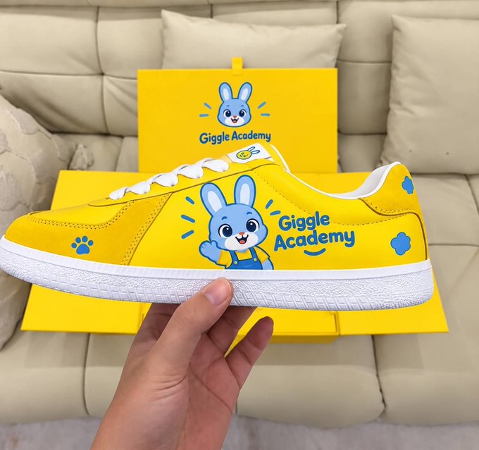A hand holds up a yellow sneaker featuring a cartoon bunny graphic and the text 'Giggle Academy', displayed in front of its matching yellow box on a beige couch.