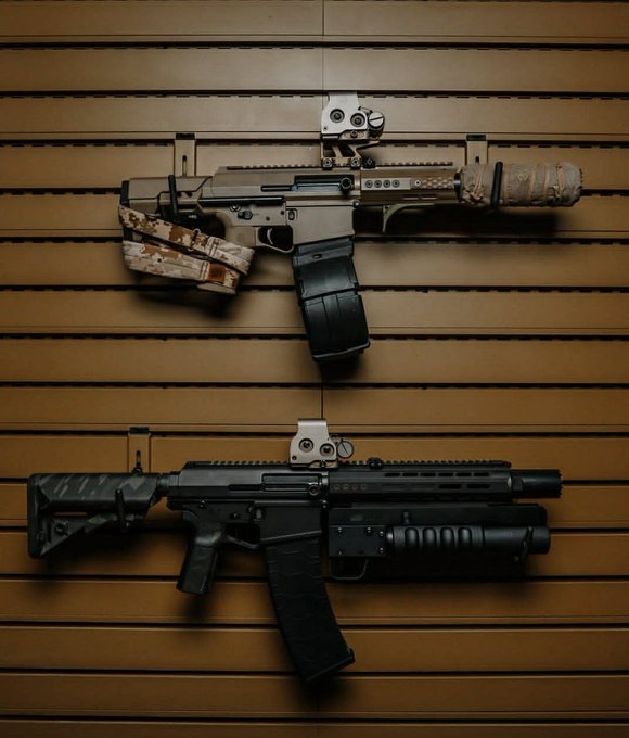 Two customized AR-style rifles are mounted on a slatwall display, the top one featuring a tan digital camouflage wrap on the stock and handguard with a suppressor and red dot sight, while the bottom one is outfitted with an underbarrel launcher, adjustable stock, and reflex optic.