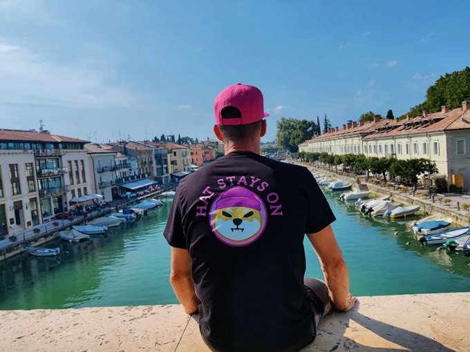 A man wearing a pink cap and a black t-shirt reading 'HAT STAYS ON' sits on a bridge overlooking a picturesque canal lined with buildings and boats.