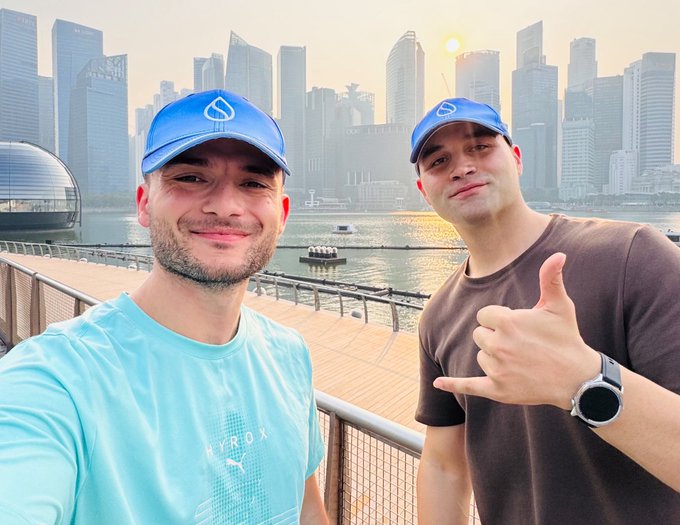 Two men pose for a selfie outdoors with a city skyline and waterfront in the background; the man on the left wears a turquoise HYROX t-shirt and a blue cap, while the man on the right wears a brown t-shirt, blue cap, and a smartwatch, giving a thumbs-up.