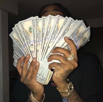 A person holds a fan of US $100 bills in front of their face, wearing multiple rings including a large diamond ring, a gold watch, and bracelets, with tattooed hands visible.