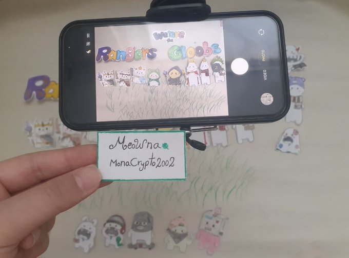 A smartphone on a tripod displaying a hand-drawn 'Rainbow Globes' NFT illustration, with a hand holding a label reading 'Moolina' and 'MonaCrypto2002' in front of it.