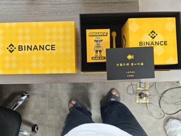 The image shows a person standing next to two boxes, one yellow and one black, with the word "BNANCE" written on them. The person appears to be wearing sandals and has their feet visible above the boxes. There are also some chairs in the background of the scene.