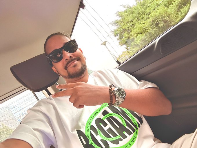 A man wearing sunglasses and a white graphic t-shirt sits in the back of a car, flashing a hand gesture toward the camera.