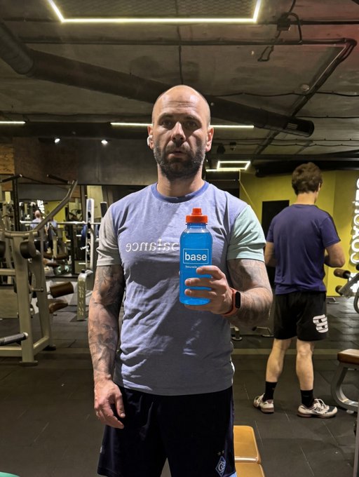 A bald, bearded, muscular man in a grey t-shirt standing in a gym holding a blue 'base' brand water bottle, with another gym-goer visible in the background.