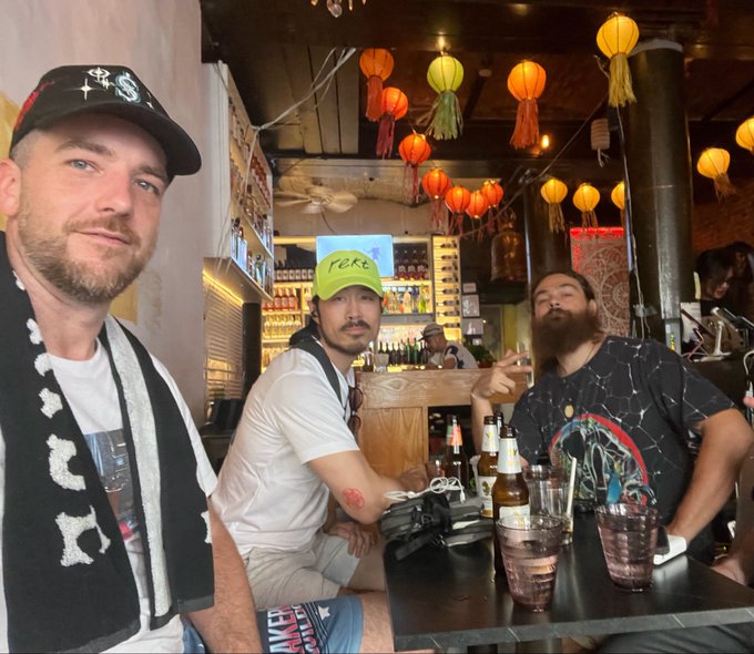Three men posing at a bar with colorful hanging lanterns, one wearing a green 'FUCK' cap and another with a full beard, drinks on the table.