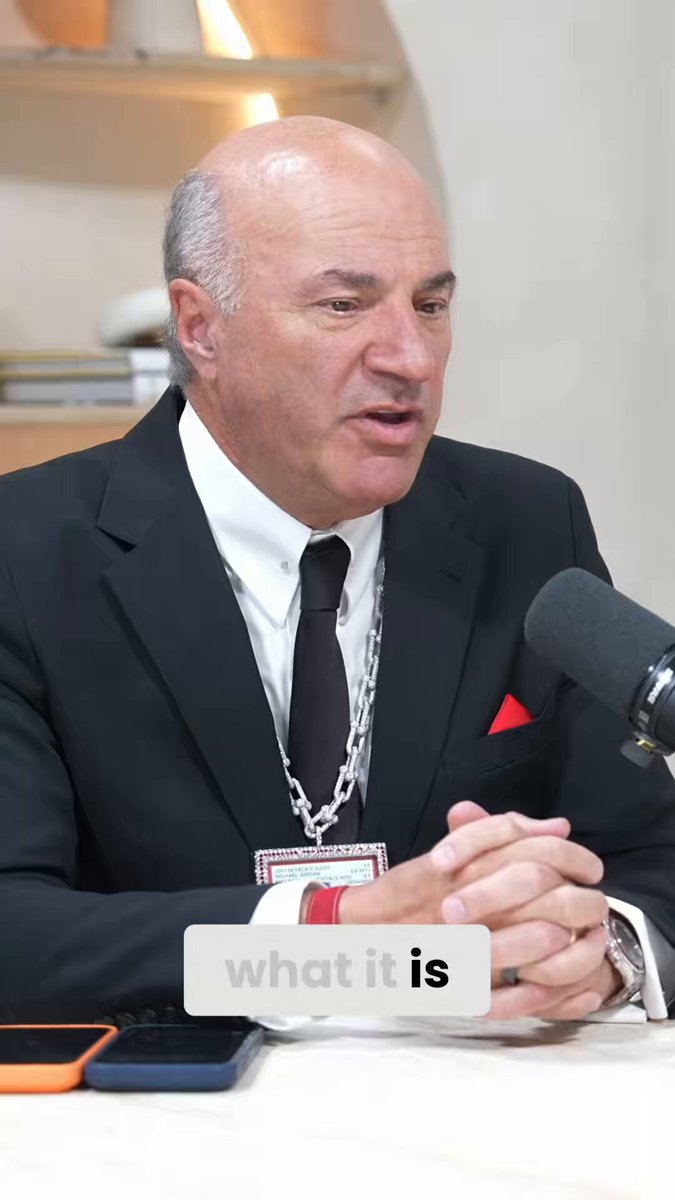 The image shows a man wearing a suit, sitting at a table with his hands folded in front of him. He has a microphone behind him, suggesting that he might be speaking or participating in an event where he needs to address an audience. The text "What it's" is visible in the image, which could possibly be related to the content being discussed by the man or the context of the situation.