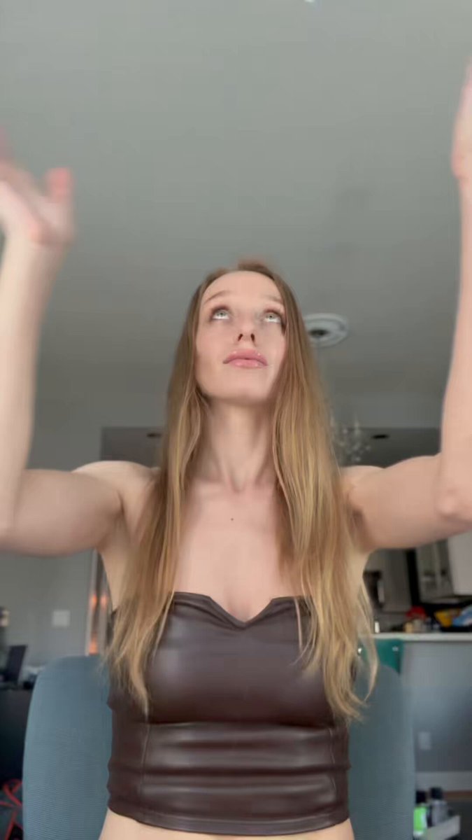 The image features a woman wearing a brown leather top, with her arms raised up in the air as if she's celebrating or expressing joy. The background shows a kitchen area with various appliances such as an oven and a microwave visible. There are also some bottles placed on the countertop near the oven. The post seems to be about a ring called the @Tangem Ring, which is being discussed by the woman in the image.