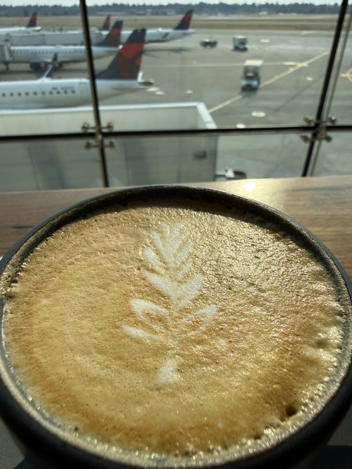 A close-up shot of a latte with foam art sitting on a wooden counter, with airplanes on a tarmac visible through the window in the background.