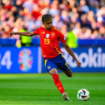 Soccer player Lamine Yamal (#19) wearing the red Spain national team jersey kicks a ball during a Euro 2024 match, with a 'Booking' sponsor sign visible behind him.